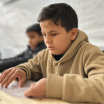 Niño con discapacidad visual en Gaza leyendo con material adaptado en un entorno educativo improvisado, símbolo de resiliencia y acceso a la educación en medio del conflicto