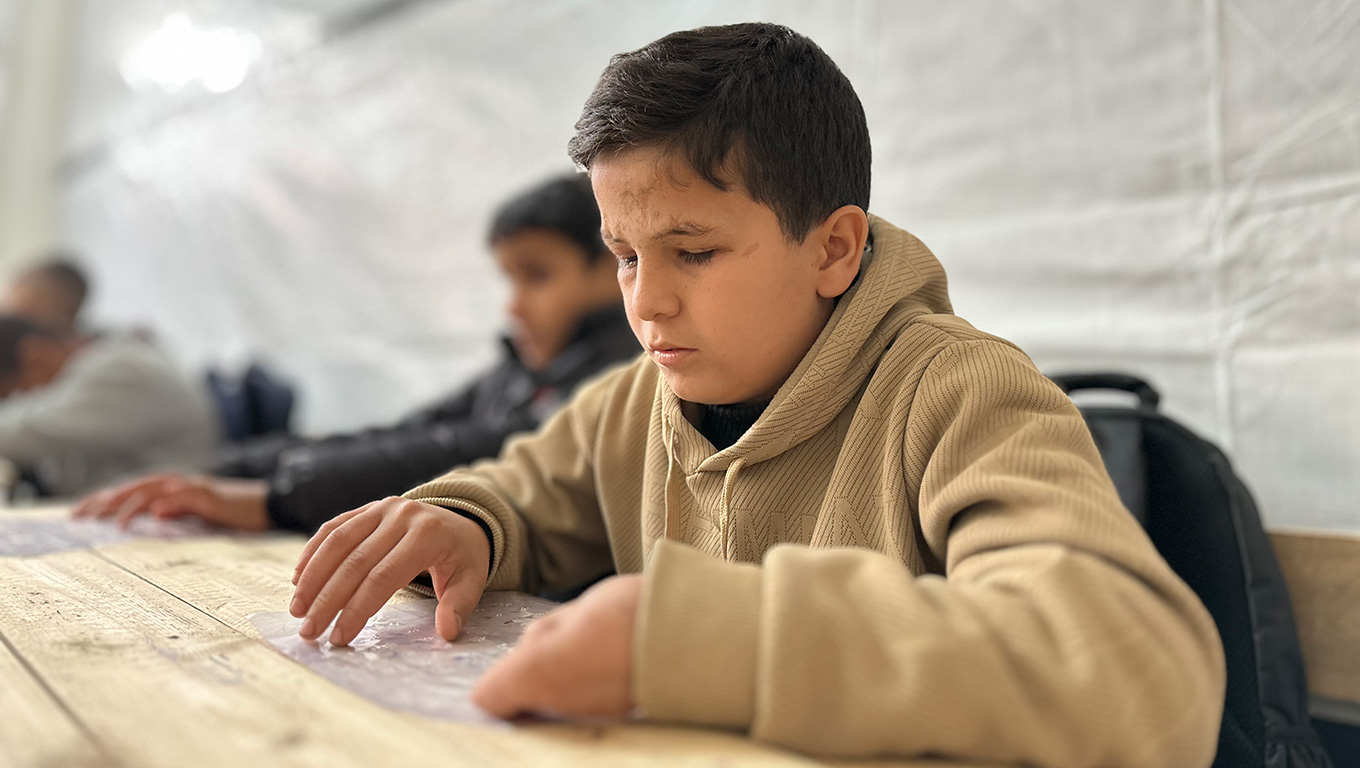 Niño con discapacidad visual en Gaza leyendo con material adaptado en un entorno educativo improvisado, símbolo de resiliencia y acceso a la educación en medio del conflicto