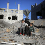 Adulto sentado junto a dos niños entre los escombros en Gaza